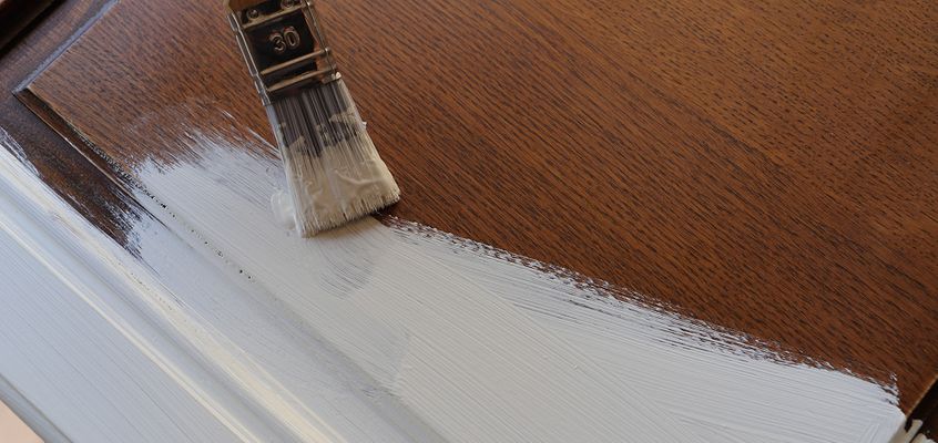 Brush applying a fresh coat of white paint over a wood cabinet door
