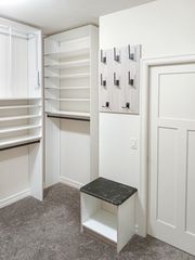 White TFL walk-in closet in Omaha with tall shoe shelves, a small bench with a dark top, and wall-mounted hooks beside the door. Custom Walk-In Closet, Omaha