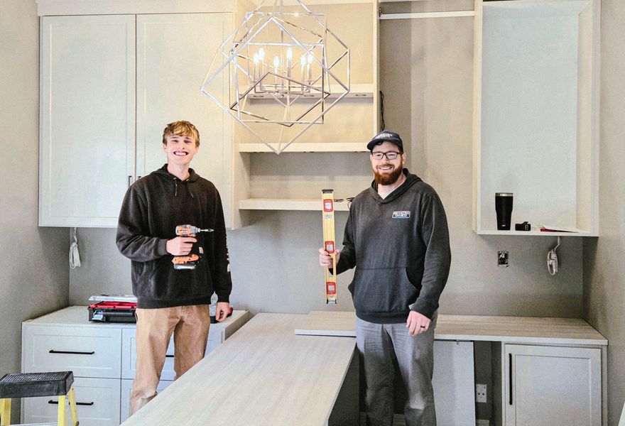 Home office installation in Omaha with light gray TFL cabinets, open shelving, and installers positioning the desktop during setup. Home Office Installation, Omaha
