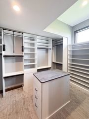 Walk-in closet in Omaha with light woodgrain finish, central island, valet rods, open shelving, and a full-height shoe wall by the window. Walk-In Closet with Island and Shoe Wall, Omaha