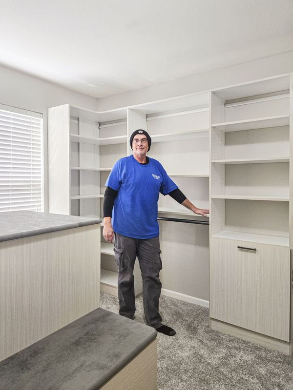 Walk-in closet installation in Omaha with light woodgrain TFL cabinets, open shelving, tall storage sections, and a gray-top island as the installer finishes setup. Walk-In Closet Installation, Omaha