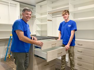 Walk-in closet installation in Omaha with light woodgrain TFL island, open shelving, and double-hang sections as installers check drawer alignment. Walk-In Closet Installation, Omaha