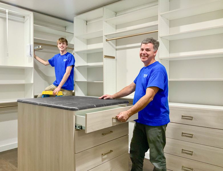 Walk-in closet installation in Omaha with light woodgrain TFL island, open shelving, double-hang sections, as installers adjust drawers. Walk-In Closet Installation, Omaha