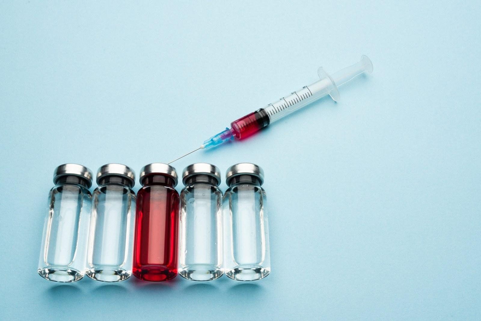 Row of glass medical vials with one red vitamin B12 vial and a syringe on a light blue background